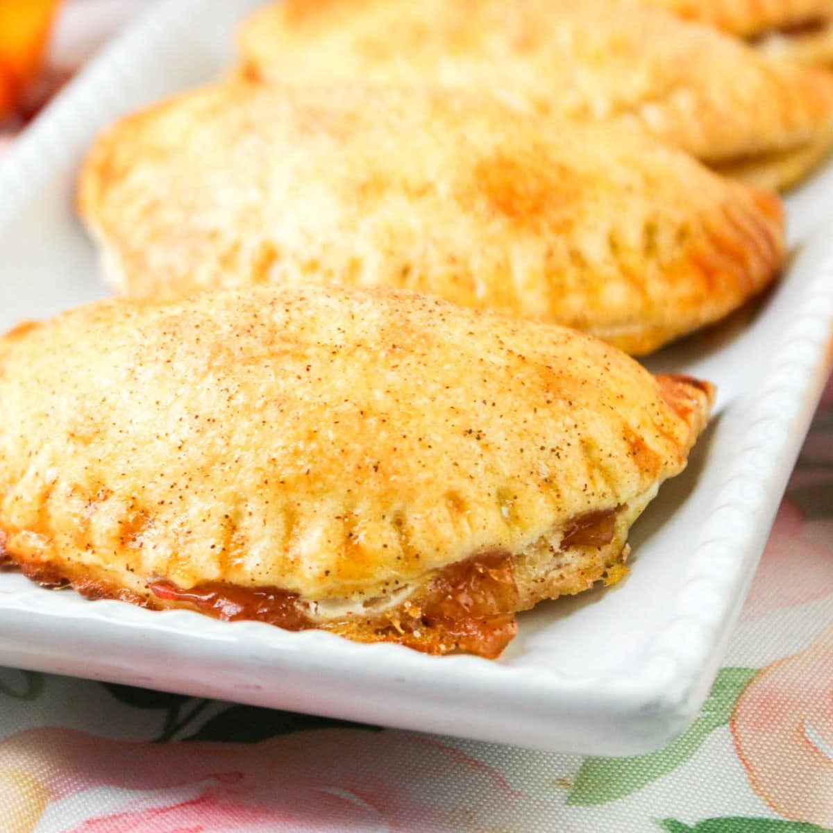 Close-up of golden-brown hand pies on a white rectangular plate, with flaky crusts and a sweet filling oozing out.