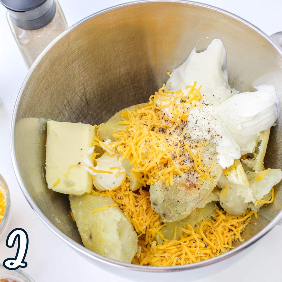 A mixing bowl with potatoes, shredded cheese, butter, sour cream, and spices. Step two of a recipe is indicated.