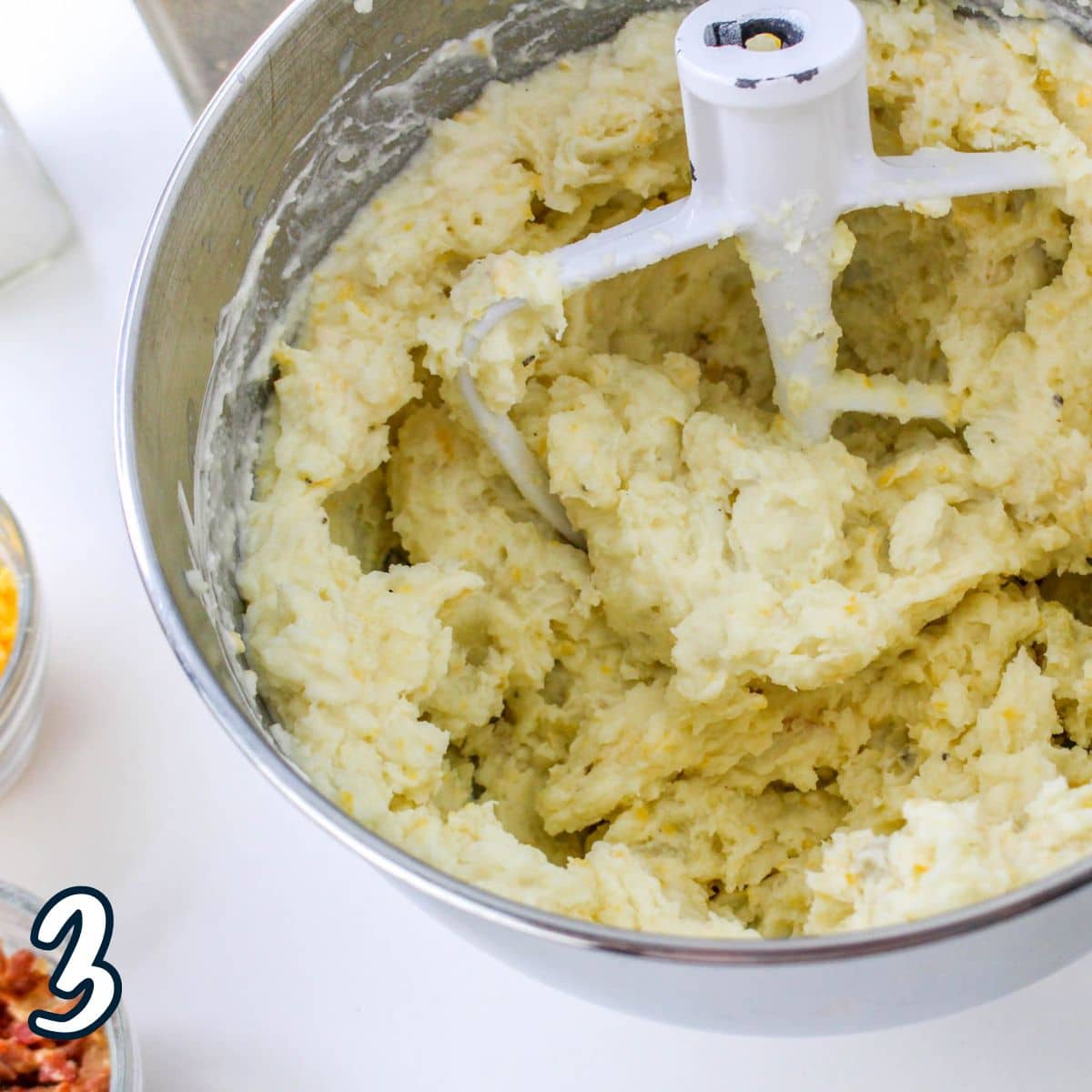 A mixing bowl with a paddle attachment, filled with creamy mashed potatoes. A number 3 is visible on the bottom left.