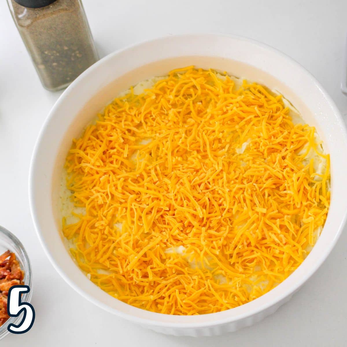 A white baking dish filled with grated cheddar cheese sits on a white surface next to seasoning containers and a bowl of bacon bits.