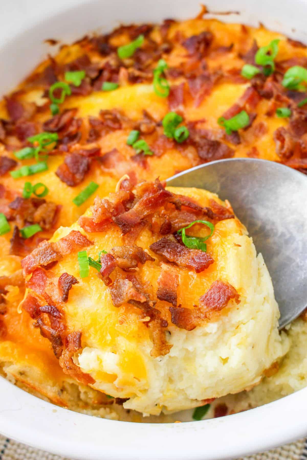 A cheesy mashed potato casserole topped with bacon bits and green onions, being served with a spoon.