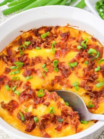 A white dish filled with baked cheesy mashed potatoes topped with bacon and green onions, with a spoon scooping a portion.