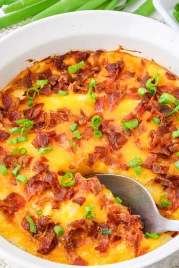 A white dish filled with baked cheesy mashed potatoes topped with bacon and green onions, with a spoon scooping a portion.