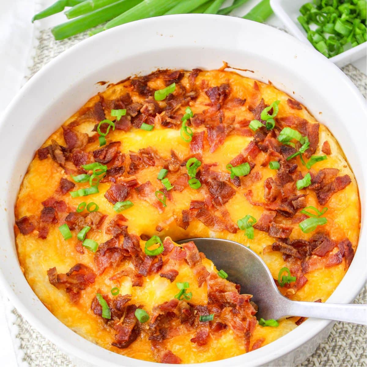 A white dish filled with baked cheesy mashed potatoes topped with bacon and green onions, with a spoon scooping a portion.