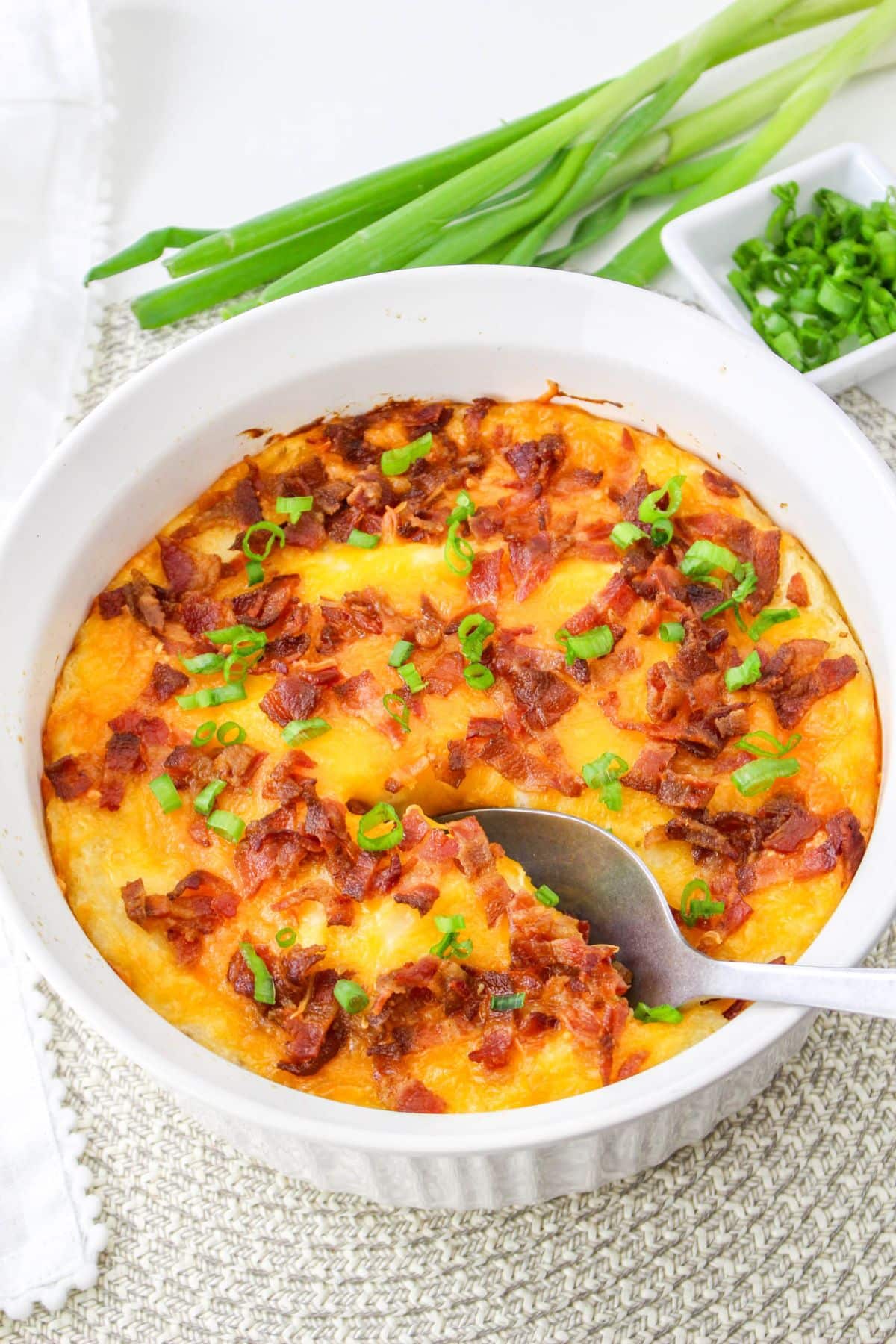 A baked casserole topped with crispy bacon bits and chopped green onions, with a spoon scooping a portion.