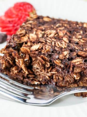 A close-up of a chocolate oatmeal with a fork, garnished with a sliced strawberry and chocolate chips on the side.