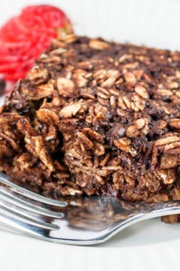 A close-up of a chocolate oatmeal with a fork, garnished with a sliced strawberry and chocolate chips on the side.