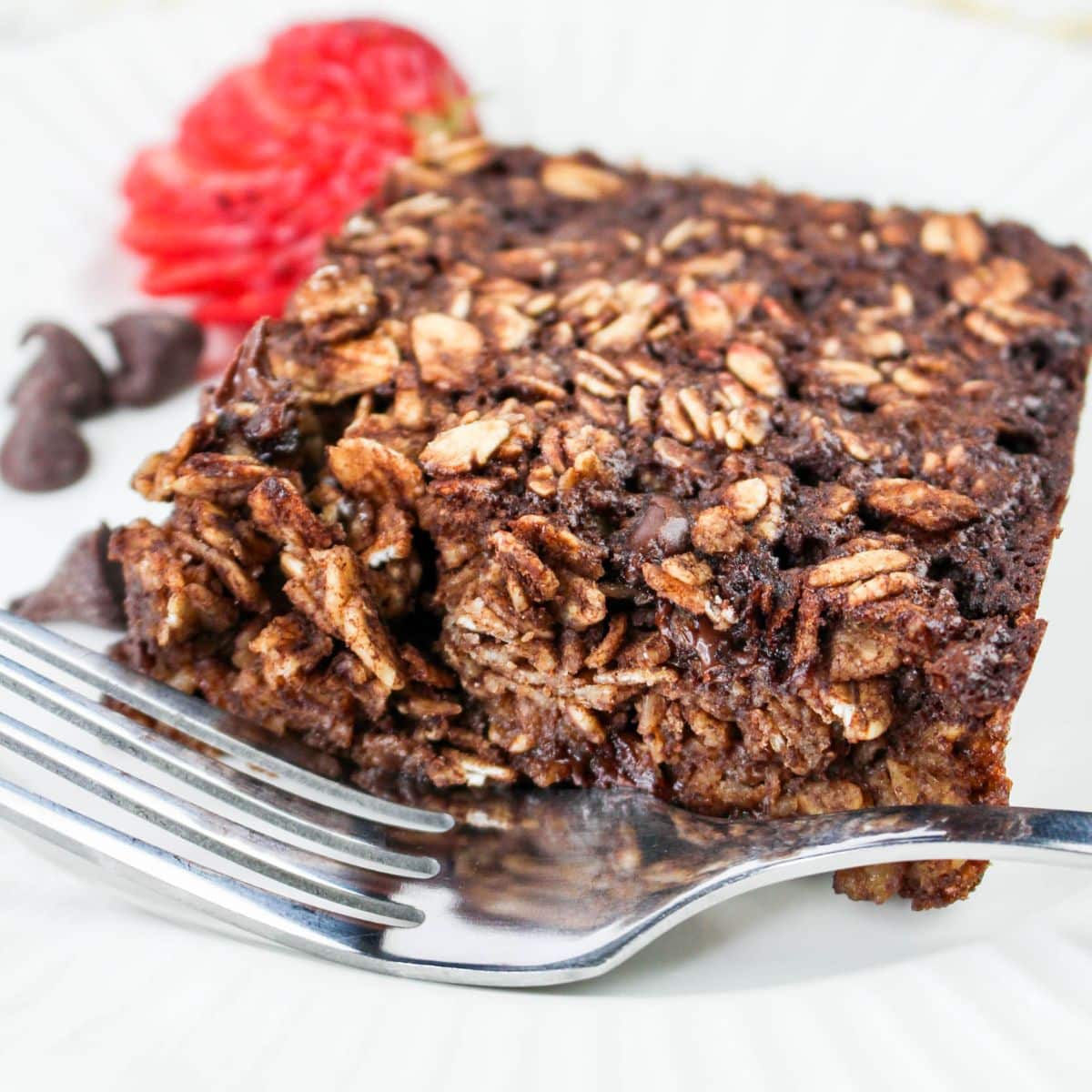 A close-up of a chocolate oatmeal with a fork, garnished with a sliced strawberry and chocolate chips on the side.