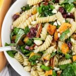 A bowl of pasta salad with spinach, roasted squash, cranberries, pecans, and feta cheese.