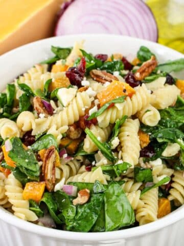 Bowl of pasta salad with spinach, pecans, cranberries, and cheese in front of onion and Parmesan.