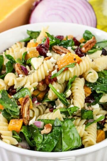Bowl of pasta salad with spinach, pecans, cranberries, and cheese in front of onion and Parmesan.