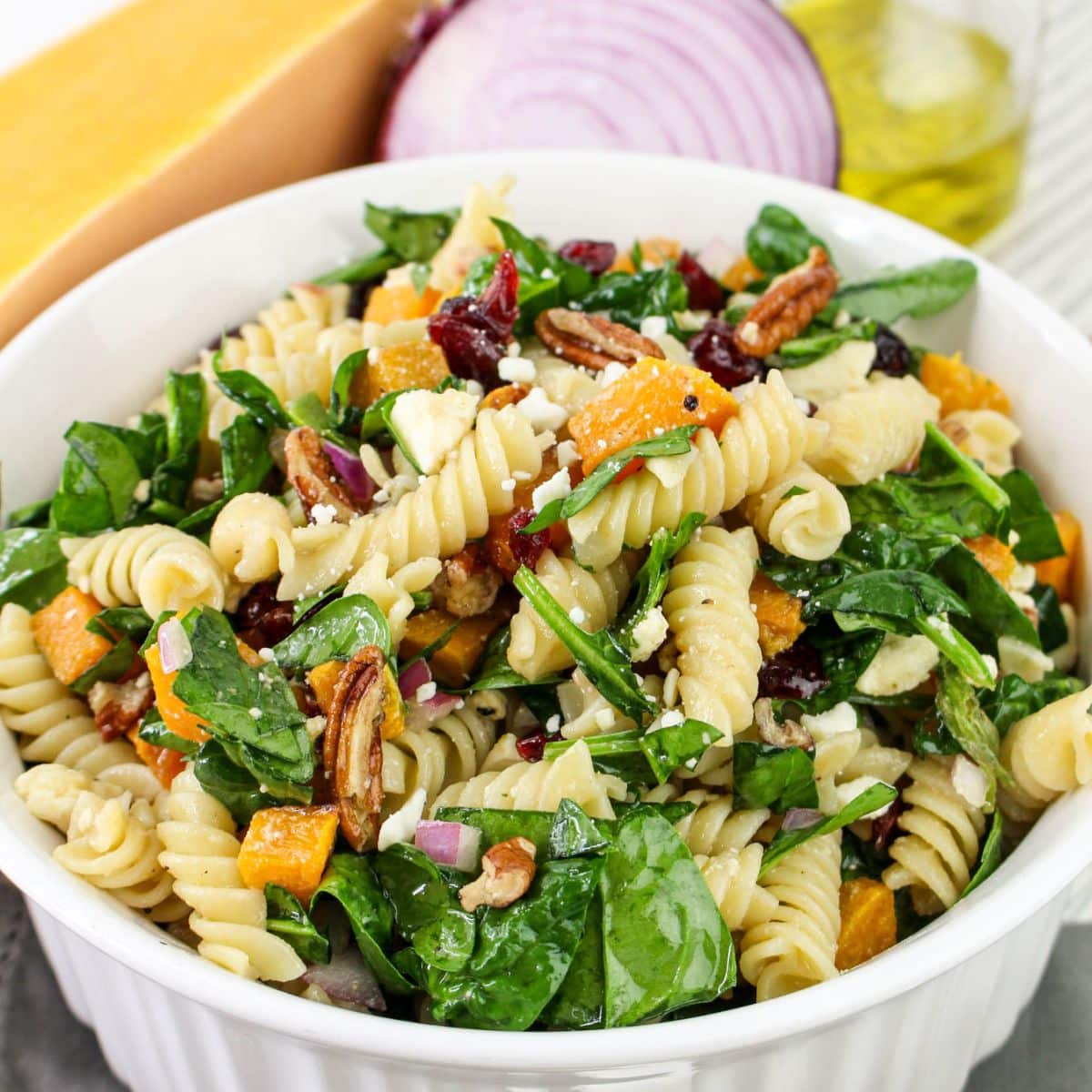 Bowl of pasta salad with spinach, pecans, cranberries, and cheese in front of onion and Parmesan.