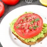 Avocado toast topped with a thick slice of tomato and sprinkled herbs on a white plate.