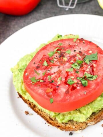 Avocado toast topped with a thick slice of tomato and sprinkled herbs on a white plate.