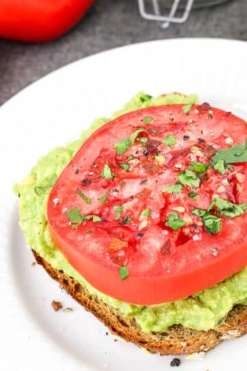 Avocado toast topped with a thick slice of tomato and sprinkled herbs on a white plate.