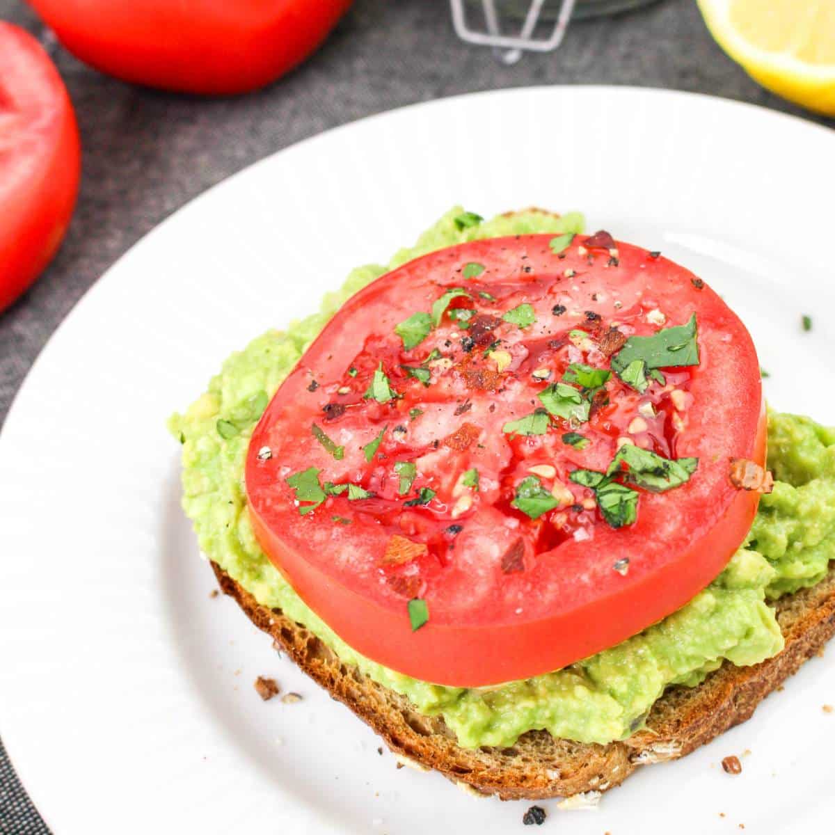 Avocado toast topped with a thick slice of tomato and sprinkled herbs on a white plate.