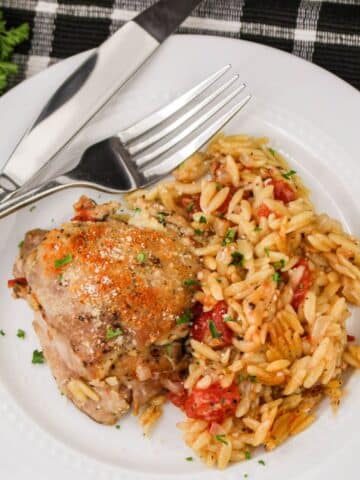 A plate of seasoned chicken with orzo pasta and tomatoes, garnished with herbs; fork and knife on the side.