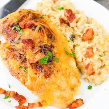 Cheesy chicken breast with bacon and herbs served alongside creamy risotto on a white plate.