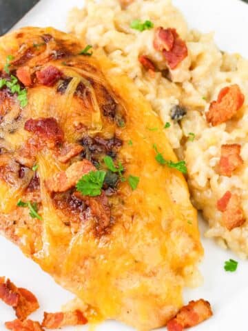 Cheesy chicken breast with bacon and herbs served alongside creamy risotto on a white plate.