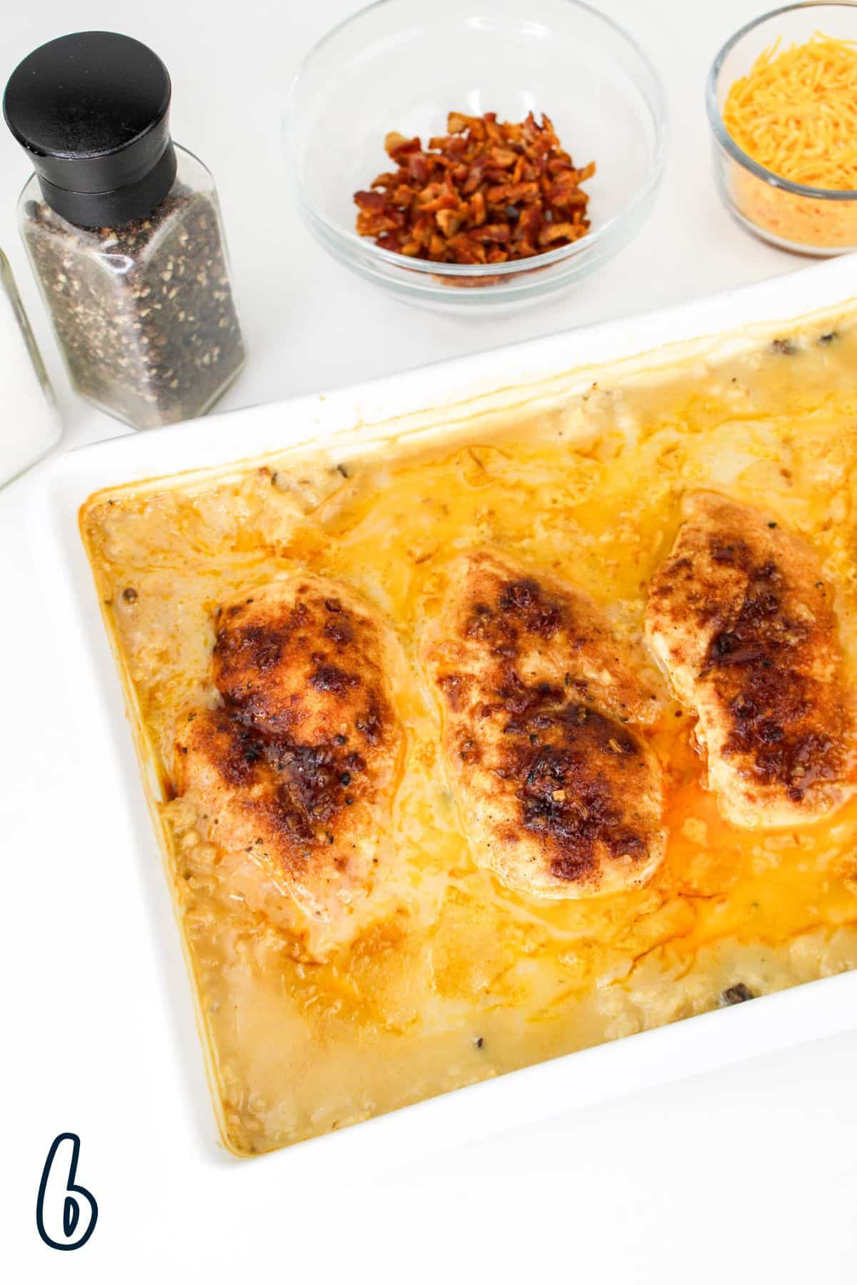 Baked chicken breasts in a dish with cheese, bacon bits, and black pepper in bowls on the side.