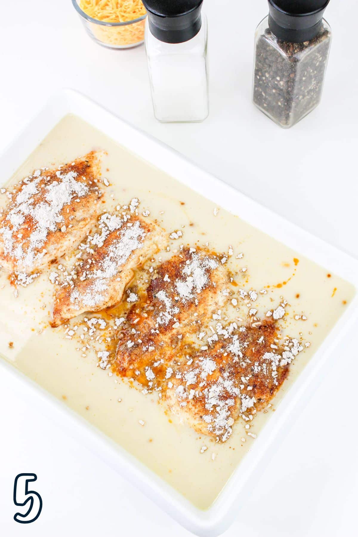 Baked chicken with sauce in a dish, topped with cheese and spices, next to salt and pepper shakers.