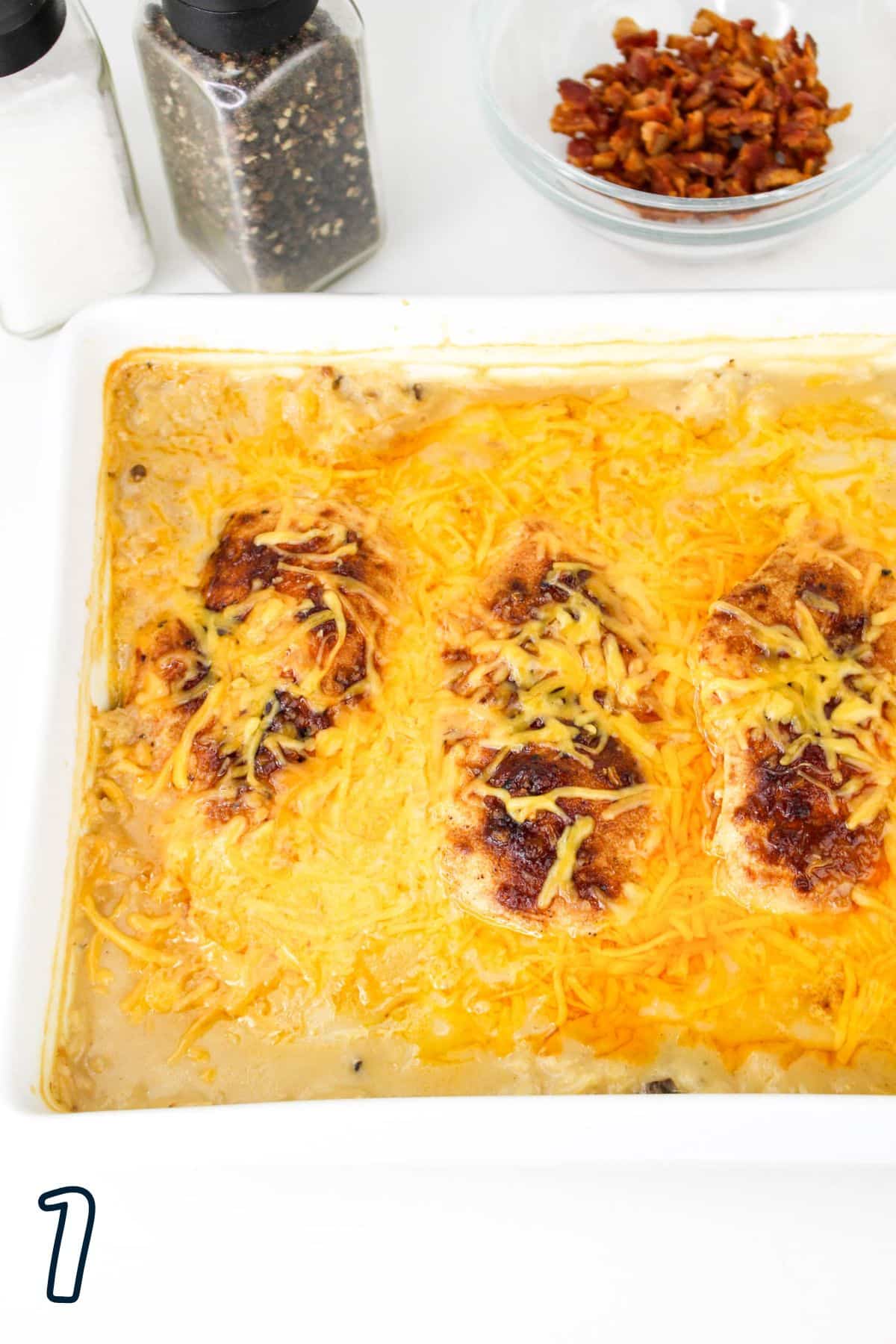 Baked chicken breasts with melted cheese in a dish, seasoning shakers, and a bowl of bacon bits nearby.