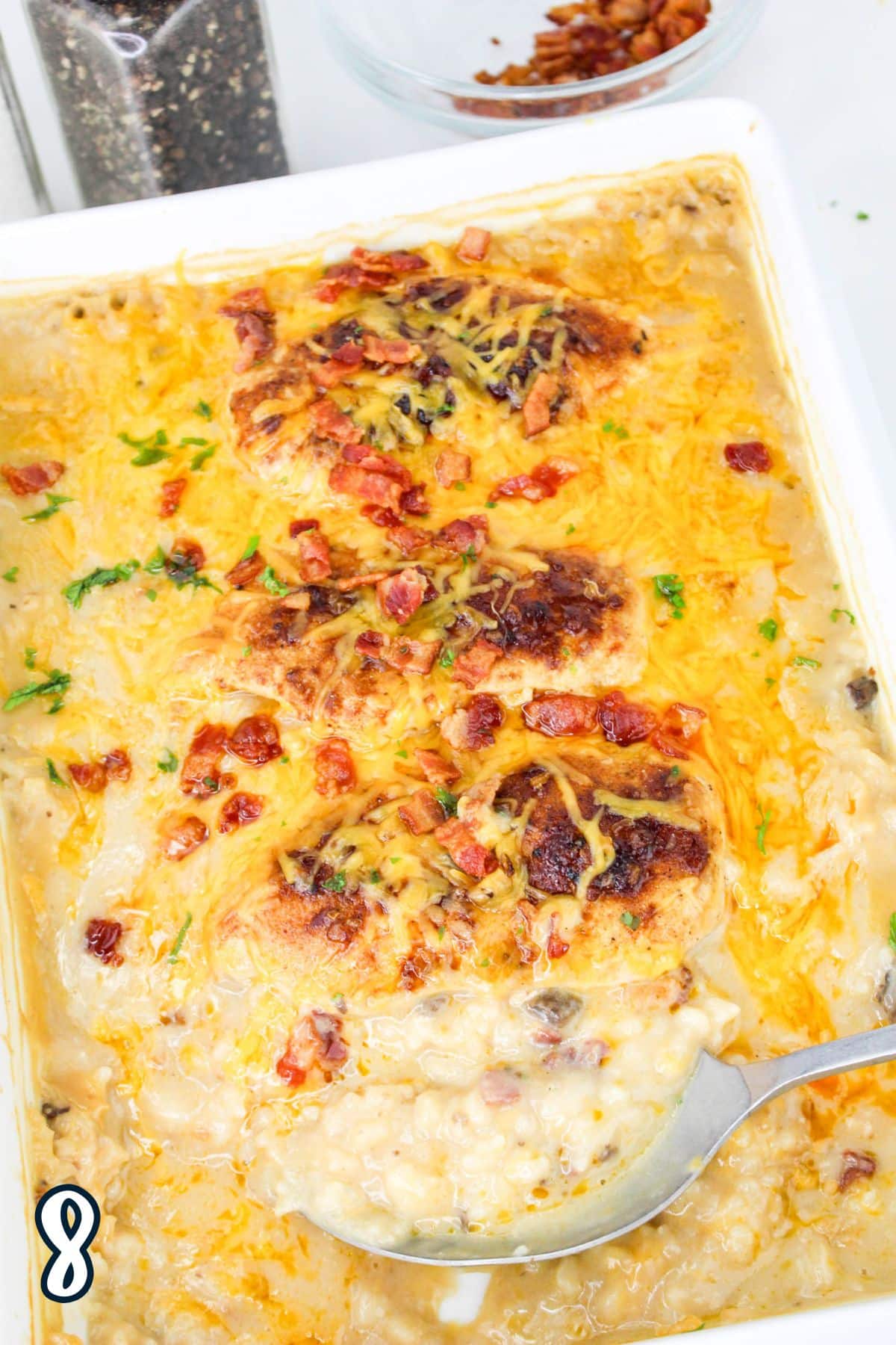 Baked chicken casserole topped with cheese and bacon in a white dish, with a serving spoon scooping it.