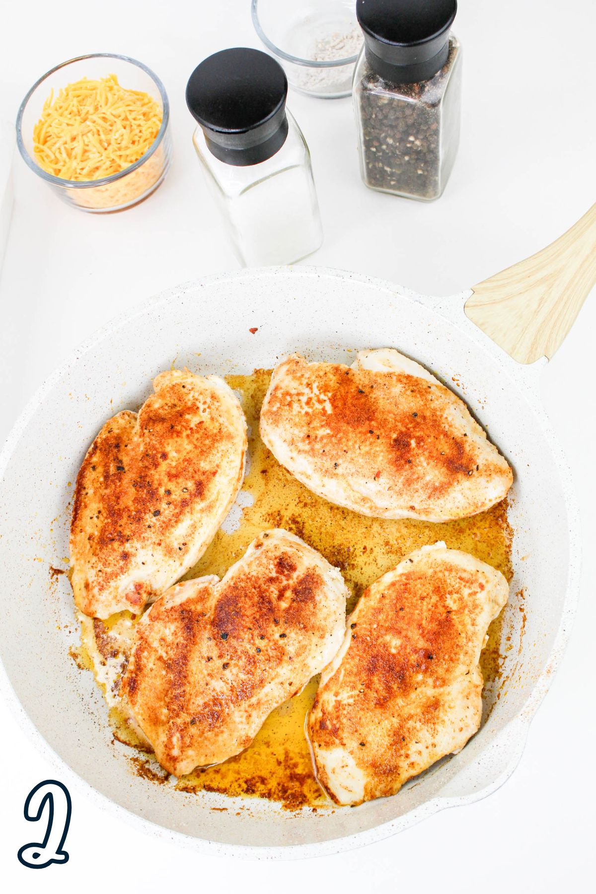 Four seasoned and cooked chicken breasts in a white pan, with spices and shredded cheese in the background.