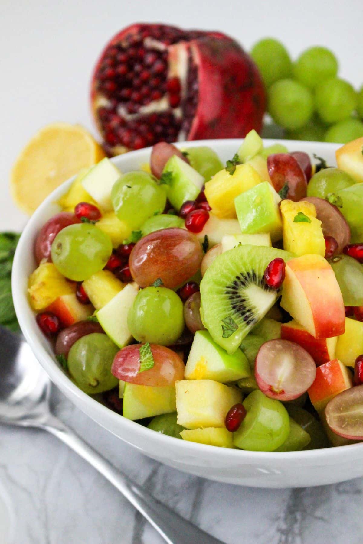 A bowl of fresh fruit salad with grapes, kiwi, apple, pineapple, and pomegranate seeds; spoon and lemons nearby.