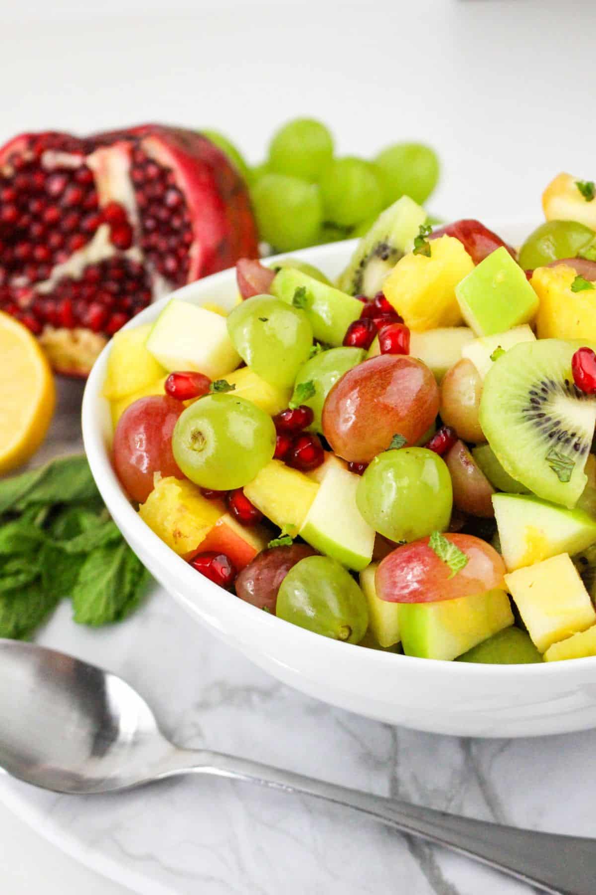 A colorful fruit salad with grapes, kiwi, pineapple, apple, and pomegranate seeds in a white bowl next to a pomegranate.