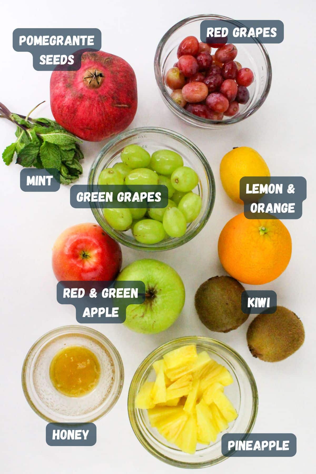 Assorted fruits in bowls: pomegranate seeds, grapes, apple, lemon, orange, kiwi, pineapple, honey, and mint.