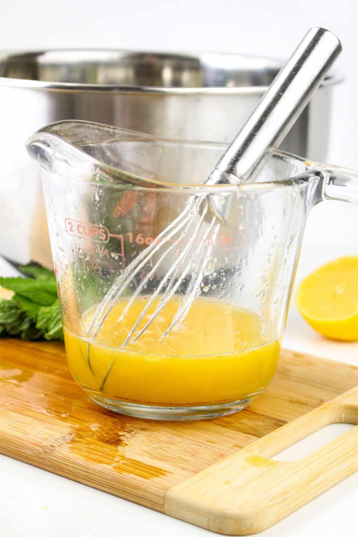 Whisk in a measuring cup with yellow liquid on a cutting board, next to half a lemon and fresh herbs.