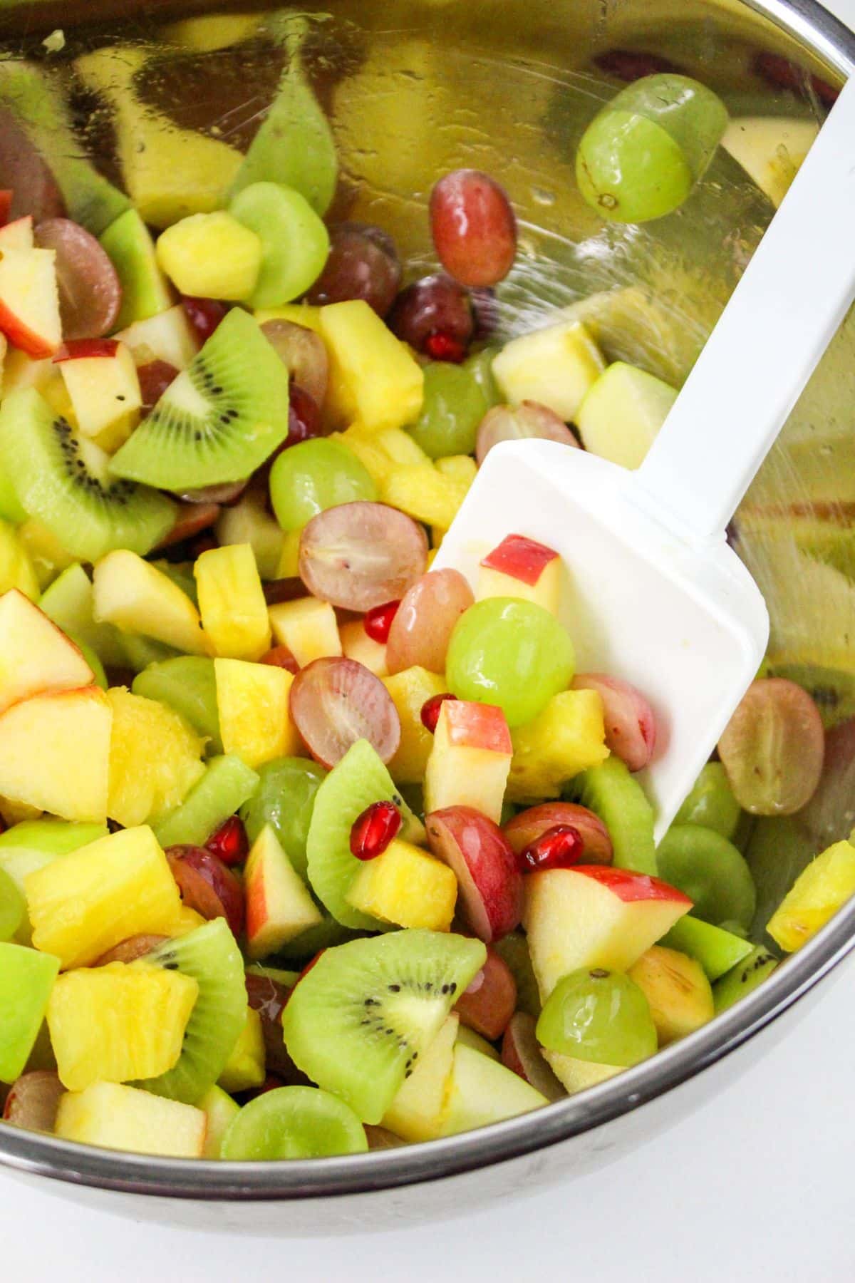 Close-up of a metal bowl with mixed fruit salad, including kiwi, grapes, apples, and pomegranate seeds, and a spatula.