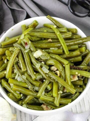 Bowl of cooked green beans with garlic and onions, placed on a gray cloth.