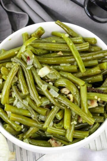 Bowl of cooked green beans with garlic and onions, placed on a gray cloth.