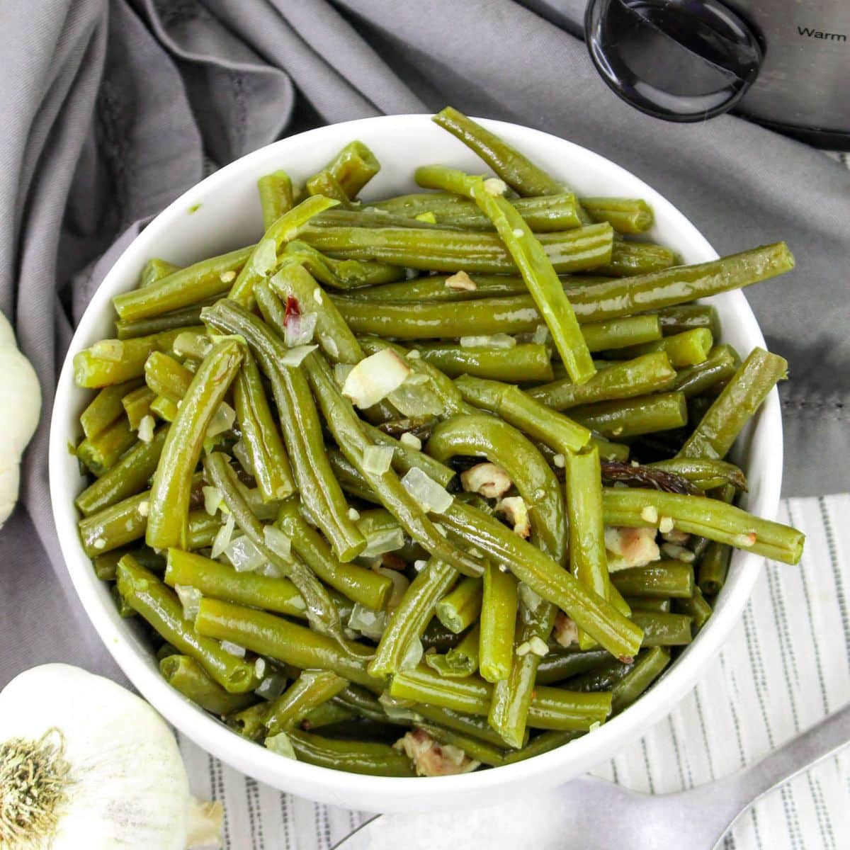 Bowl of cooked green beans with garlic and onions, placed on a gray cloth.