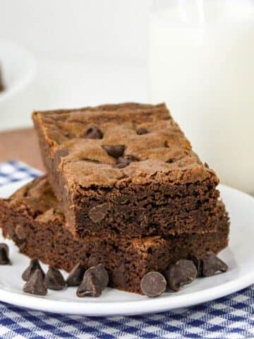 Two chocolate chip brownie cookies on a plate with chocolate chips and a glass of milk.