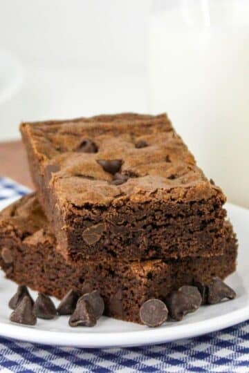 Two chocolate chip brownie cookies on a plate with chocolate chips and a glass of milk.