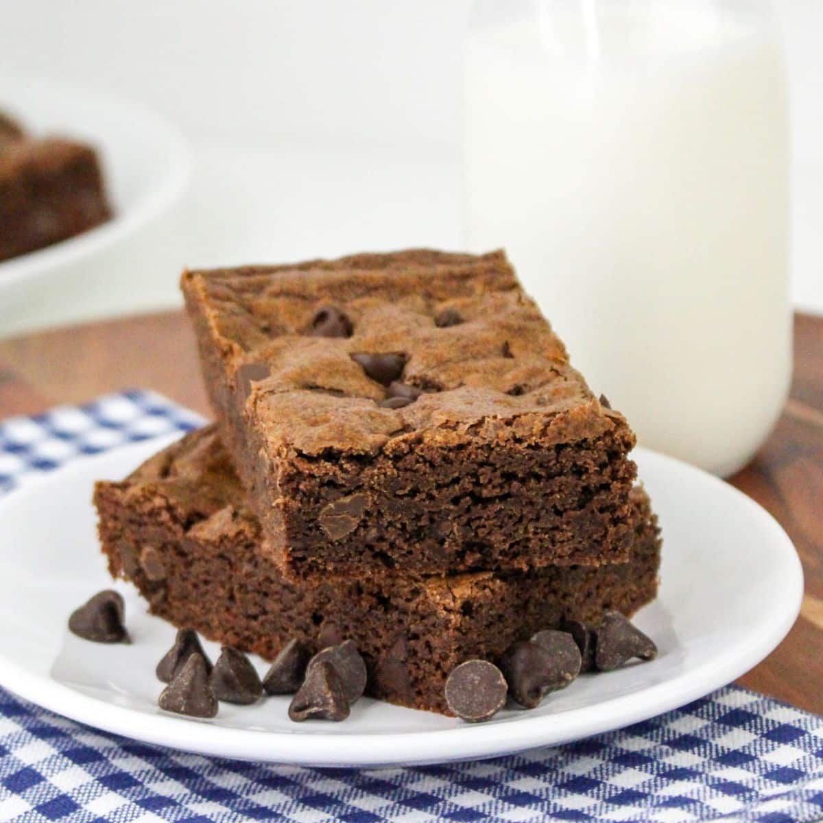 Two chocolate chip brownie cookies on a plate with chocolate chips and a glass of milk.