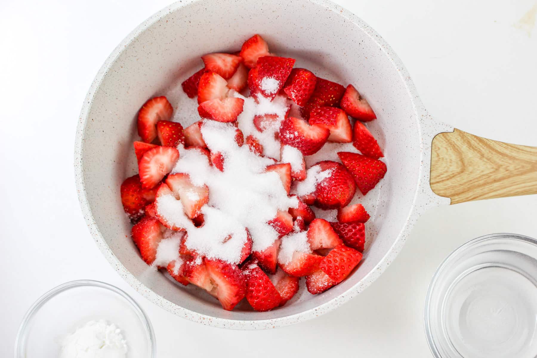 Chopped strawberries with sugar in a white saucepan, surrounded by a small glass bowl, evoke memories of a delightful chocolate covered strawberry cake.