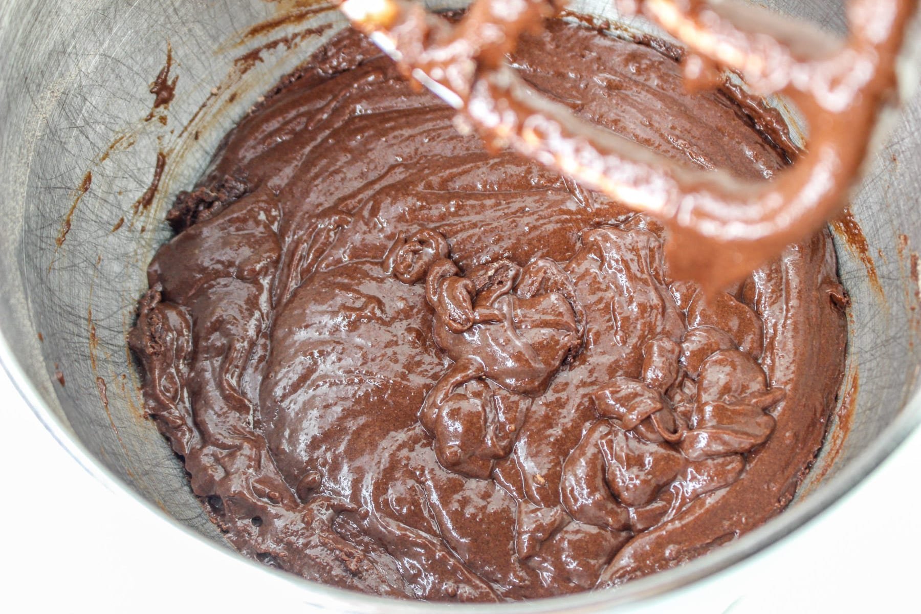 Thick, rich chocolate batter in a metal mixing bowl with part of a beater showing, promising the indulgence of a chocolate-covered strawberry cake.