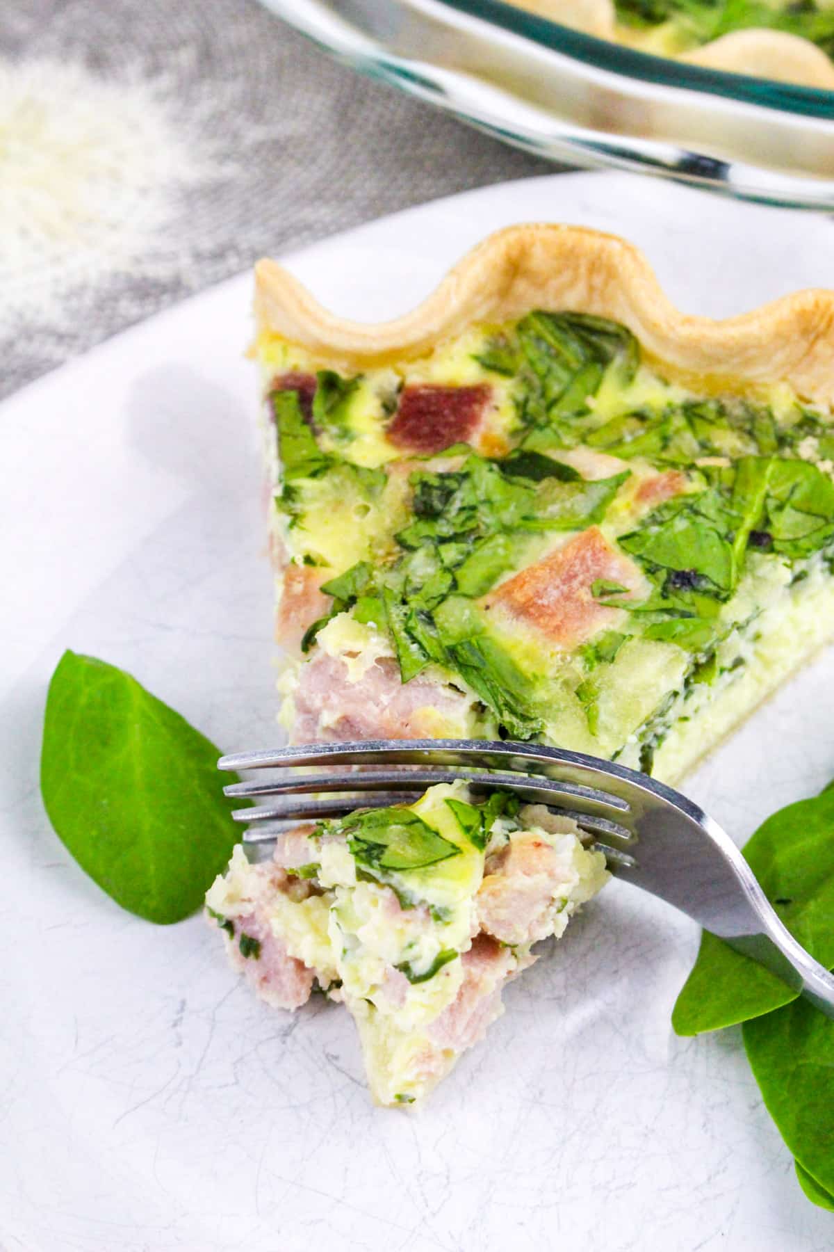 A slice of quiche with spinach and ham on a plate, garnished with spinach leaves, and a fork taking a bite.