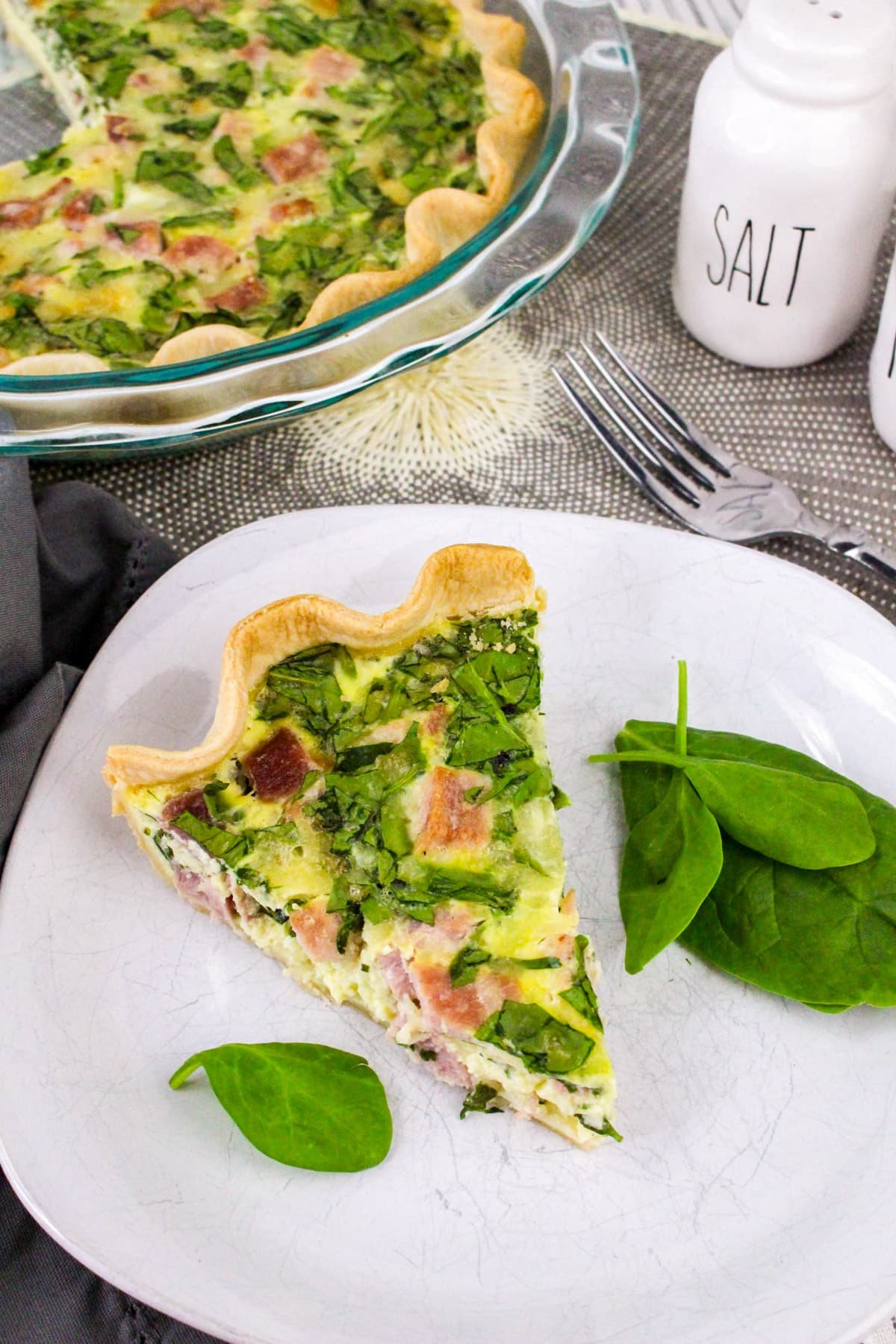 A slice of spinach and ham quiche on a plate with fresh spinach leaves, next to a salt shaker and fork.