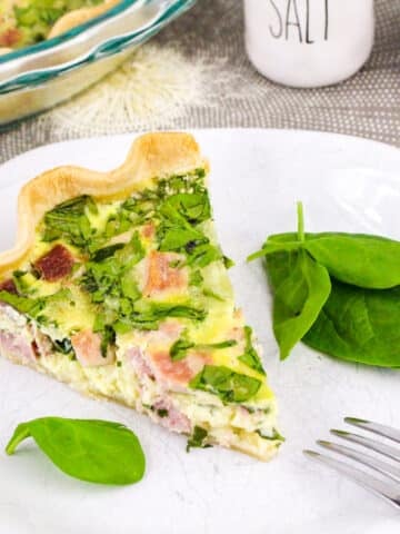 Slice of ham and spinach quiche on a plate with spinach leaves, set on a gray tablecloth next to salt and pepper shakers.