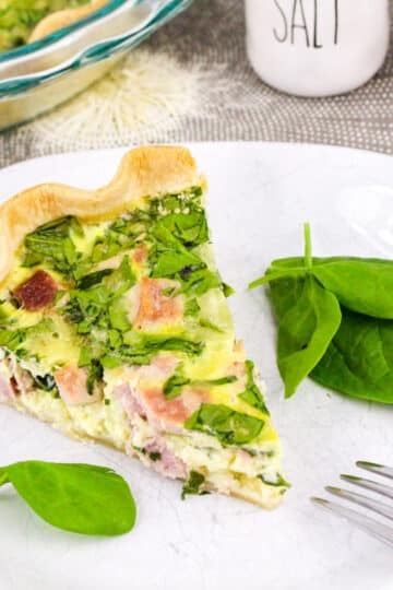 Slice of ham and spinach quiche on a plate with spinach leaves, set on a gray tablecloth next to salt and pepper shakers.