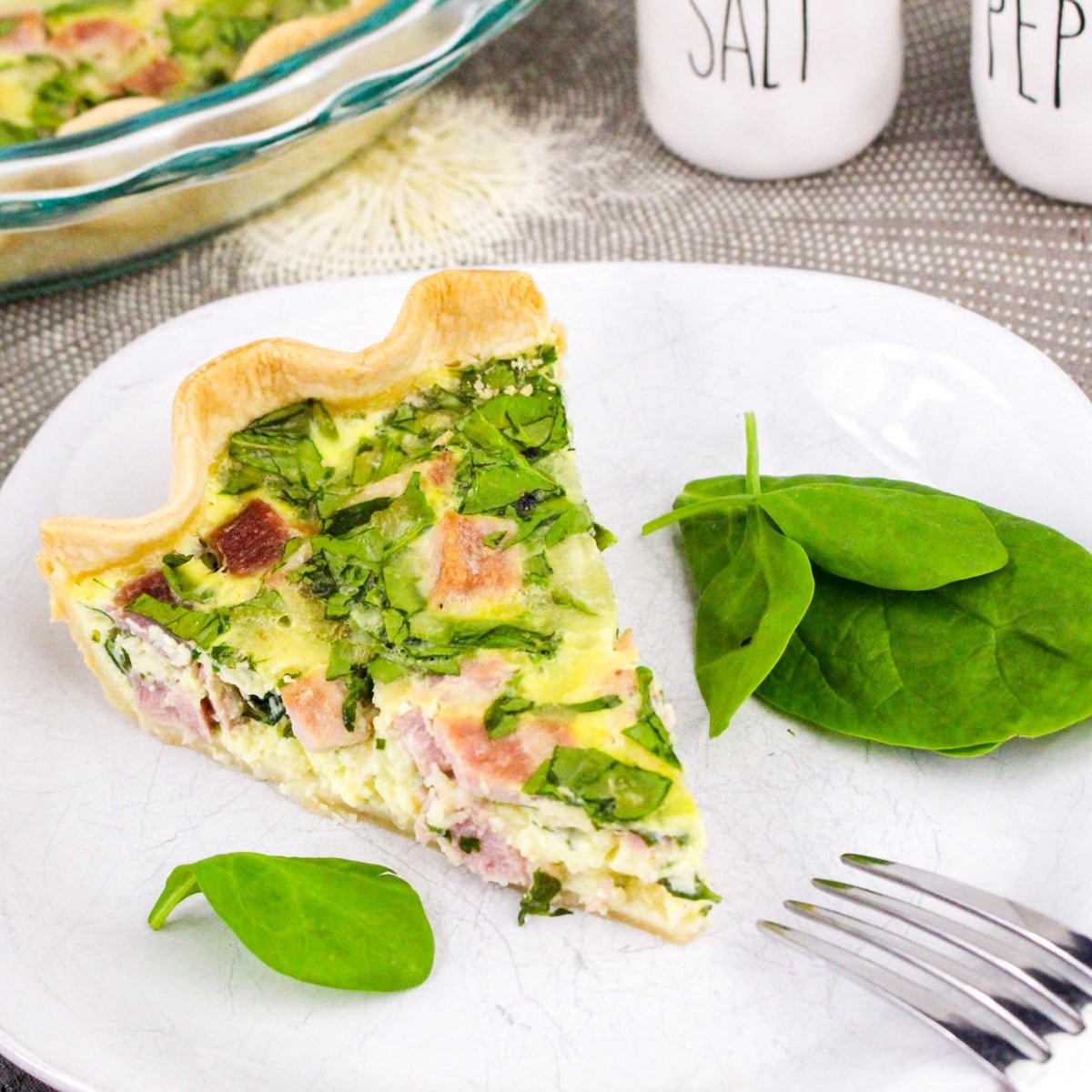 Slice of ham and spinach quiche on a plate with spinach leaves, set on a gray tablecloth next to salt and pepper shakers.