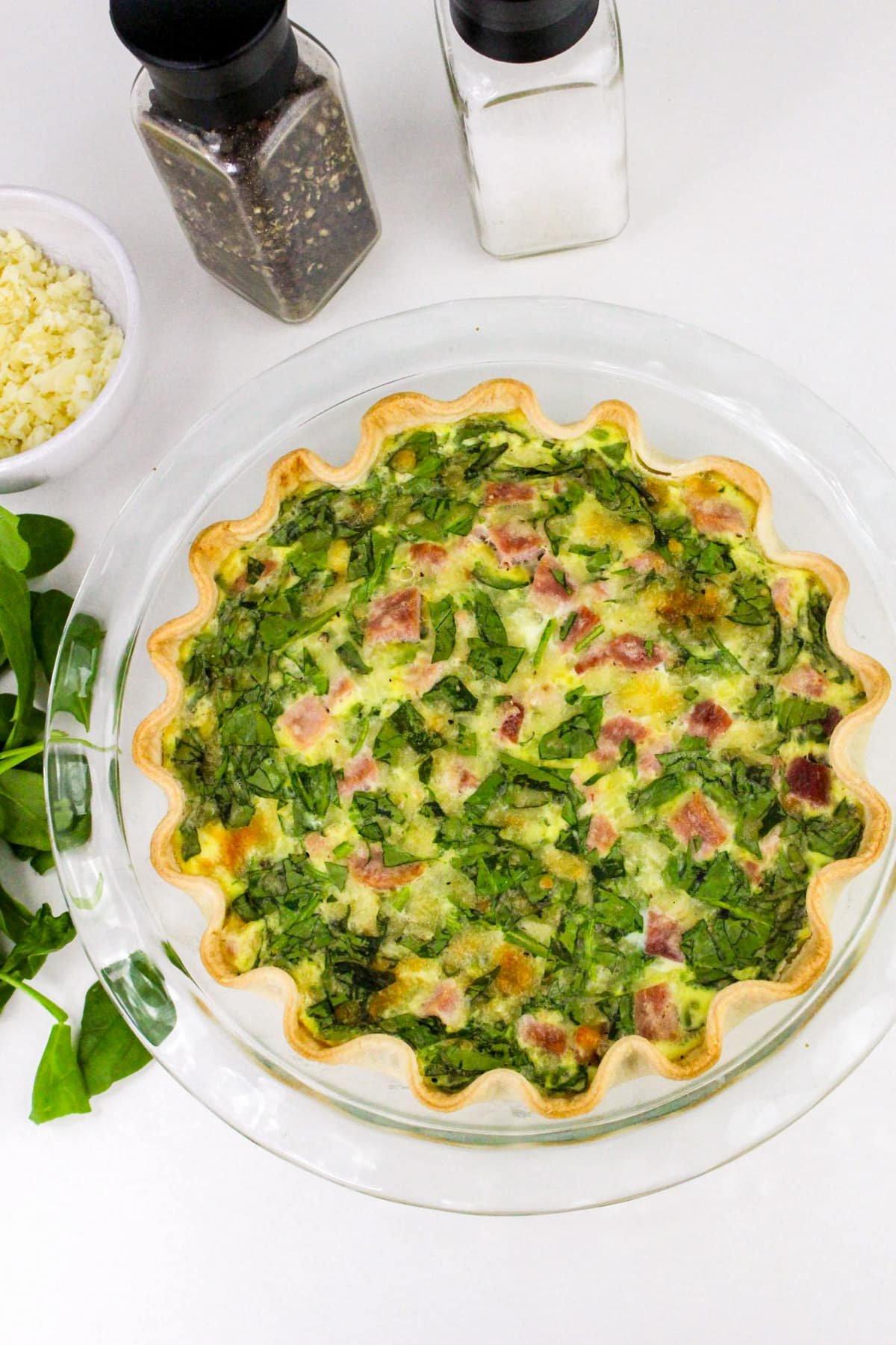 Quiche with spinach and ham in a glass pie dish, surrounded by salt, pepper, cheese, and spinach leaves.