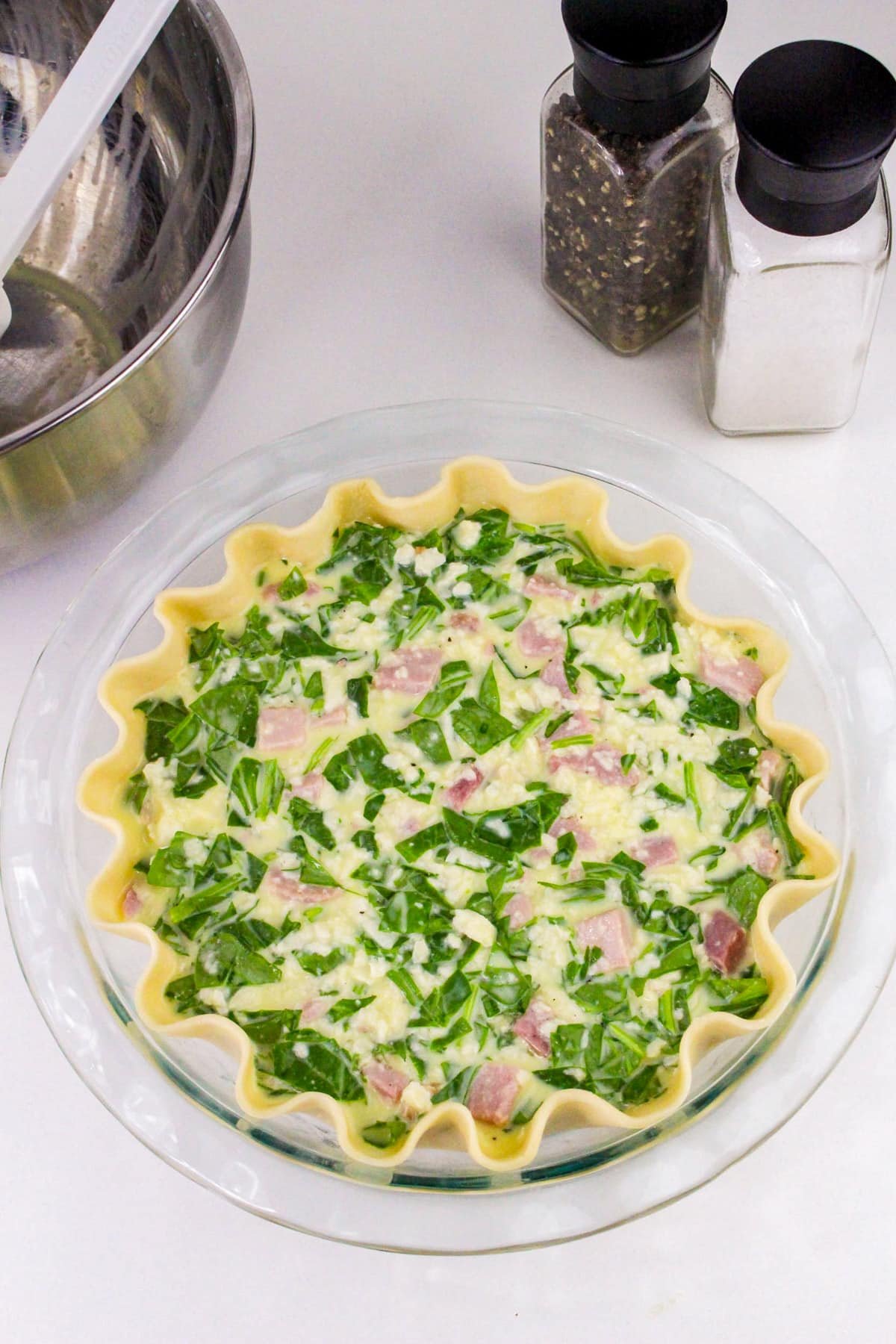 Unbaked quiche with spinach and ham in a pie crust, next to salt and pepper shakers on a white countertop.