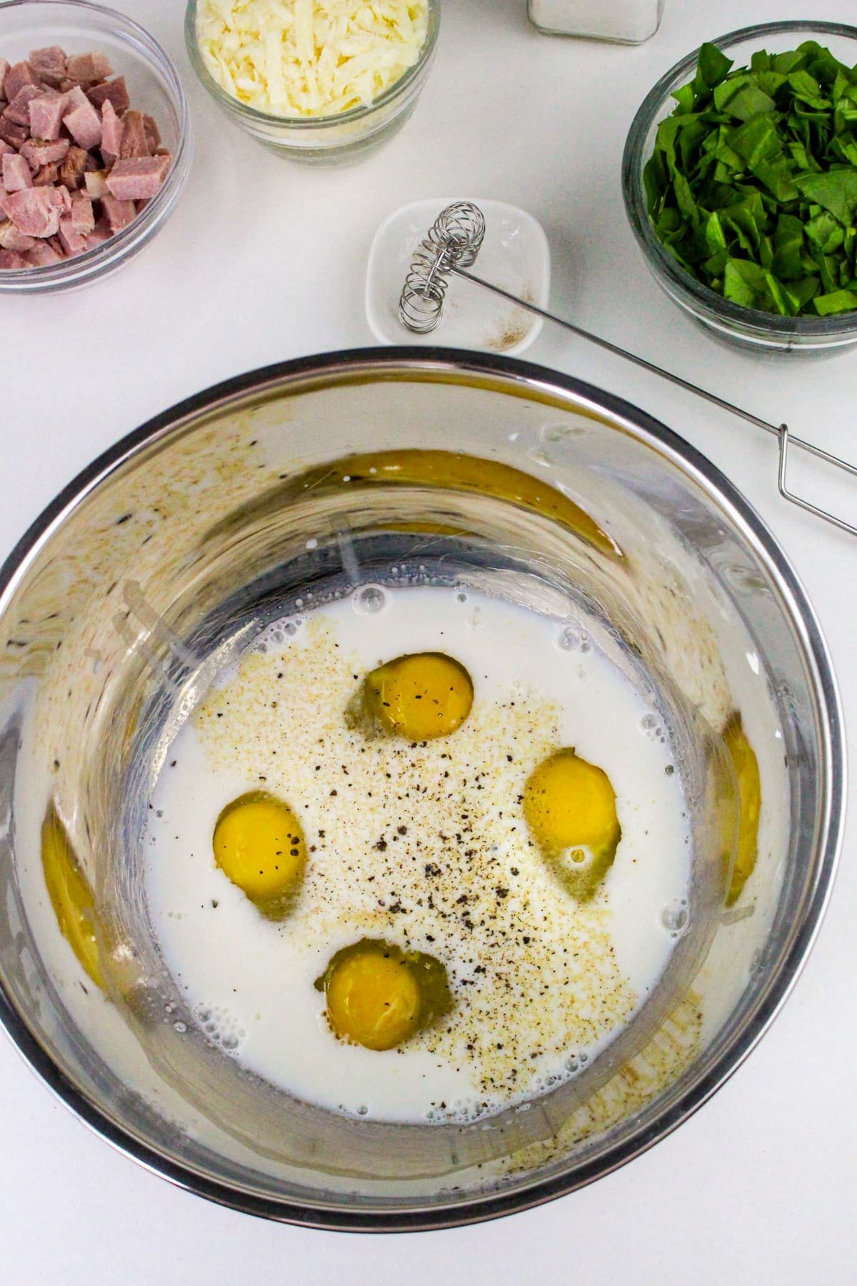 Eggs, cream, and pepper in a mixing bowl, with spinach, ham, and cheese in bowls nearby on a white countertop.