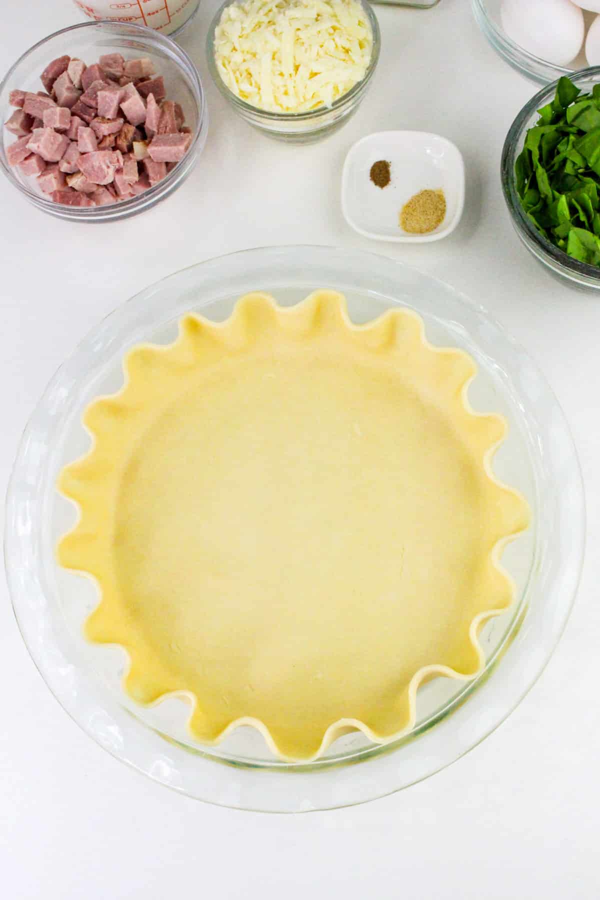 Pie crust in a glass dish surrounded by ingredients like ham, cheese, eggs, spinach, and spices on a white surface.